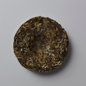 A round compressed cake of loose-leaf tea, likely Pu'er, showing a textured blend of dark brown, olive green, and light tan leaves, photographed from directly above on a neutral gray background.