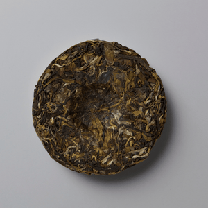 A round compressed cake of loose-leaf tea, likely Pu'er, showing a textured blend of dark brown, olive green, and light tan leaves, photographed from directly above on a neutral gray background.