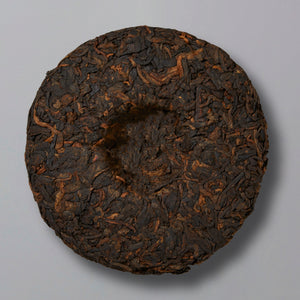 A round, compressed tea cake with various shades of dark brown and black leaves, creating a textured surface, placed on a plain light gray background.