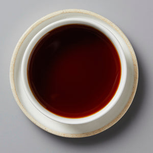 Top-down view of dark, rich brewed shou pu’er tea in a white ceramic cup and saucer on a gray surface.