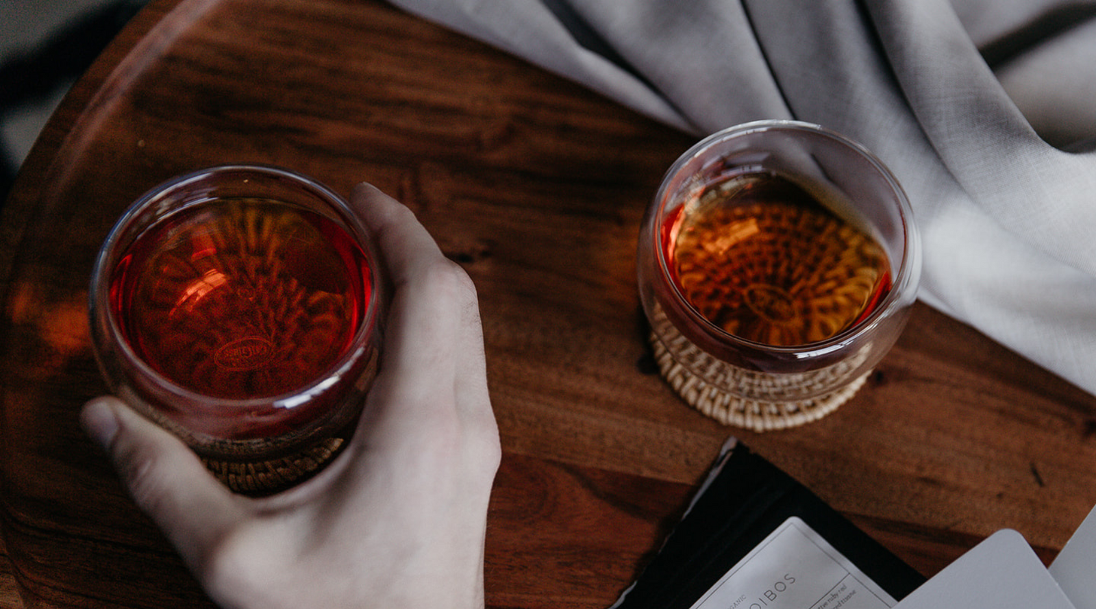 A cozy scene with two glasses of dark amber liquid on a wooden table. A hand holds one glass. A soft gray fabric drapes in the background, creating a warm, relaxing atmosphere.