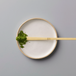 Matcha Kame ceremonial grade powder with bamboo whisk on white ceramic plate against grey background