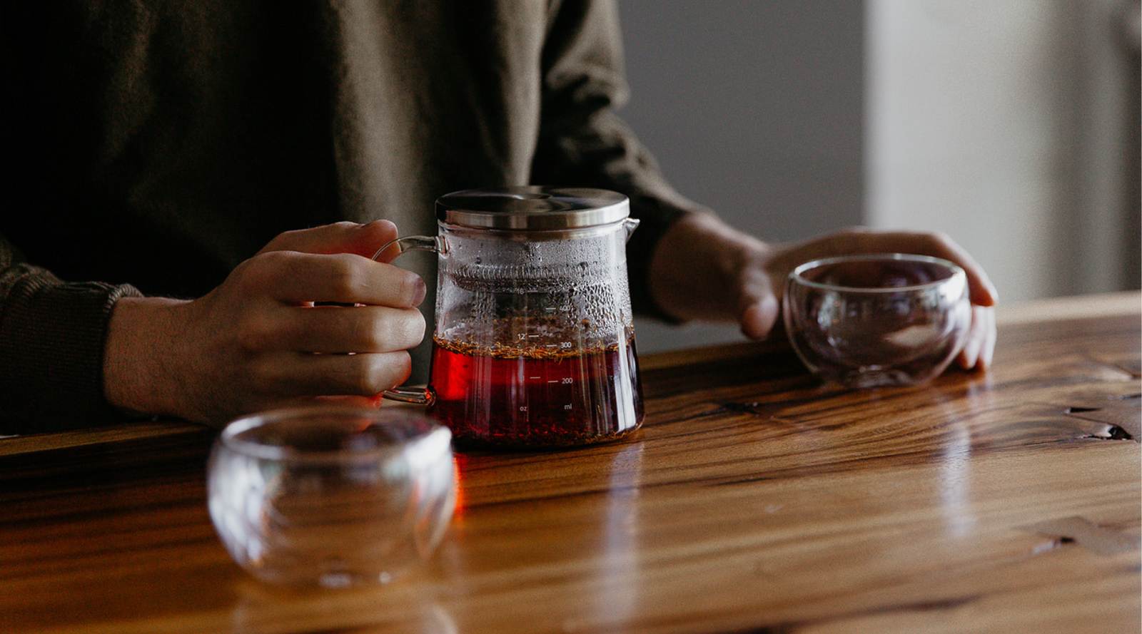 A person in a brown sweater holds a glass teapot with amber tea on a wooden table. Two empty glass teacups are nearby, creating a warm, inviting atmosphere.