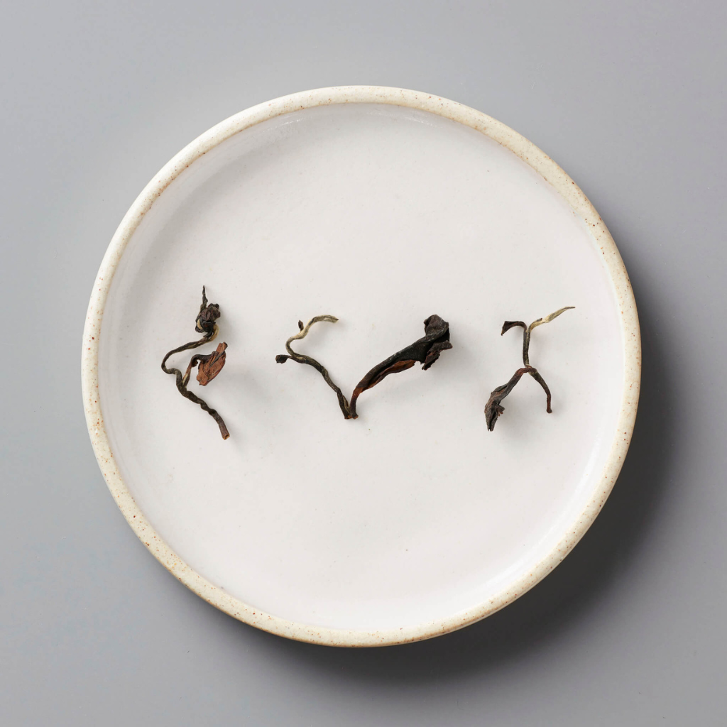 A white plate on a gray surface holds four dried tea leaves spaced apart. The muted tones suggest simplicity and focus on the texture of the leaves.