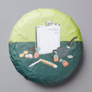 Grandpas Flask huangpian puer tea cake with illustrated wrapper showing flask, pipe and tea accessories on green background
