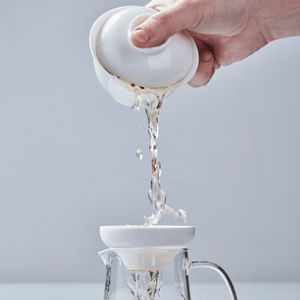 White porcelain filter pouring tea through strainer into glass pitcher, water streaming through perforated base
