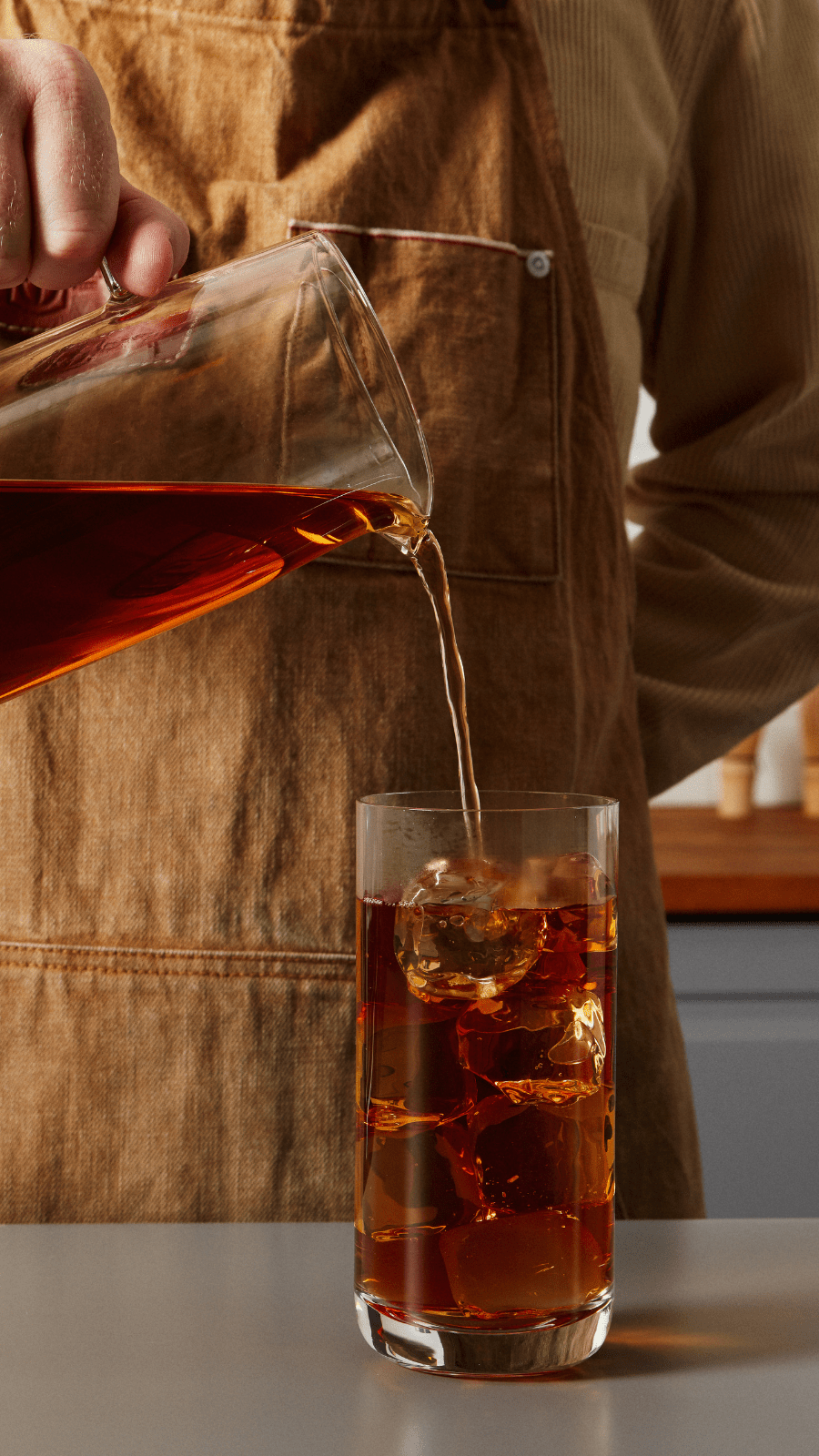 A person prepares tea in a traditional ceremony, pouring loose tea leaves from a bamboo scoop into a white gaiwan on a wooden tea tray. The setup includes white cups and a brown napkin, conveying a calm, ritualistic atmosphere.