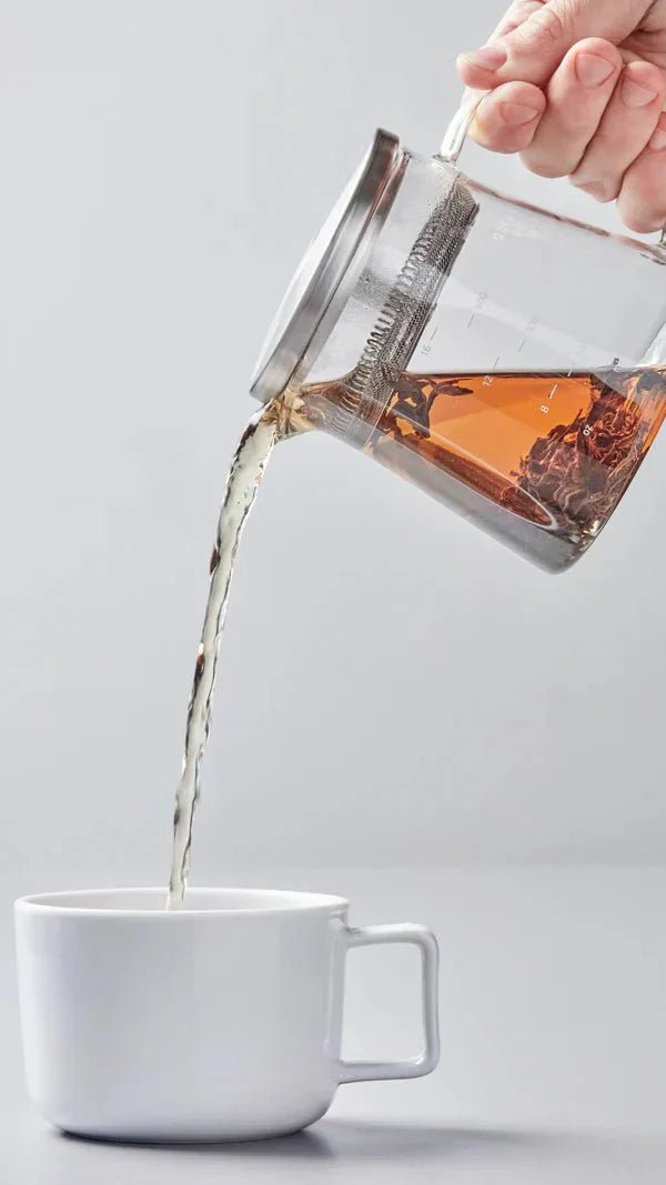 A person prepares tea in a traditional ceremony, pouring loose tea leaves from a bamboo scoop into a white gaiwan on a wooden tea tray. The setup includes white cups and a brown napkin, conveying a calm, ritualistic atmosphere.