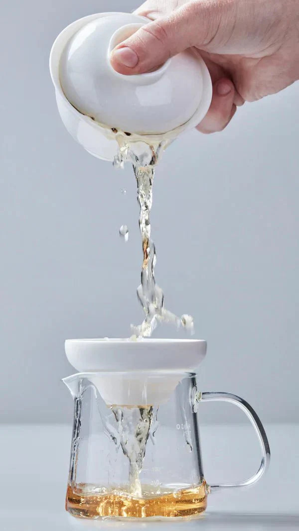 A person prepares tea in a traditional ceremony, pouring loose tea leaves from a bamboo scoop into a white gaiwan on a wooden tea tray. The setup includes white cups and a brown napkin, conveying a calm, ritualistic atmosphere.