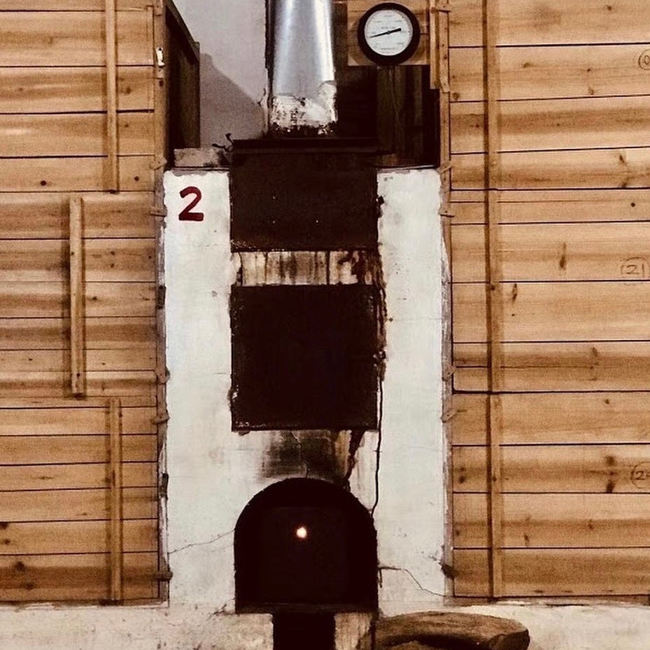 A rustic industrial oven with a metal chimney and gauge. Wooden panels surround it, and a small flame glows in the dark opening below. Number "2" is visible.