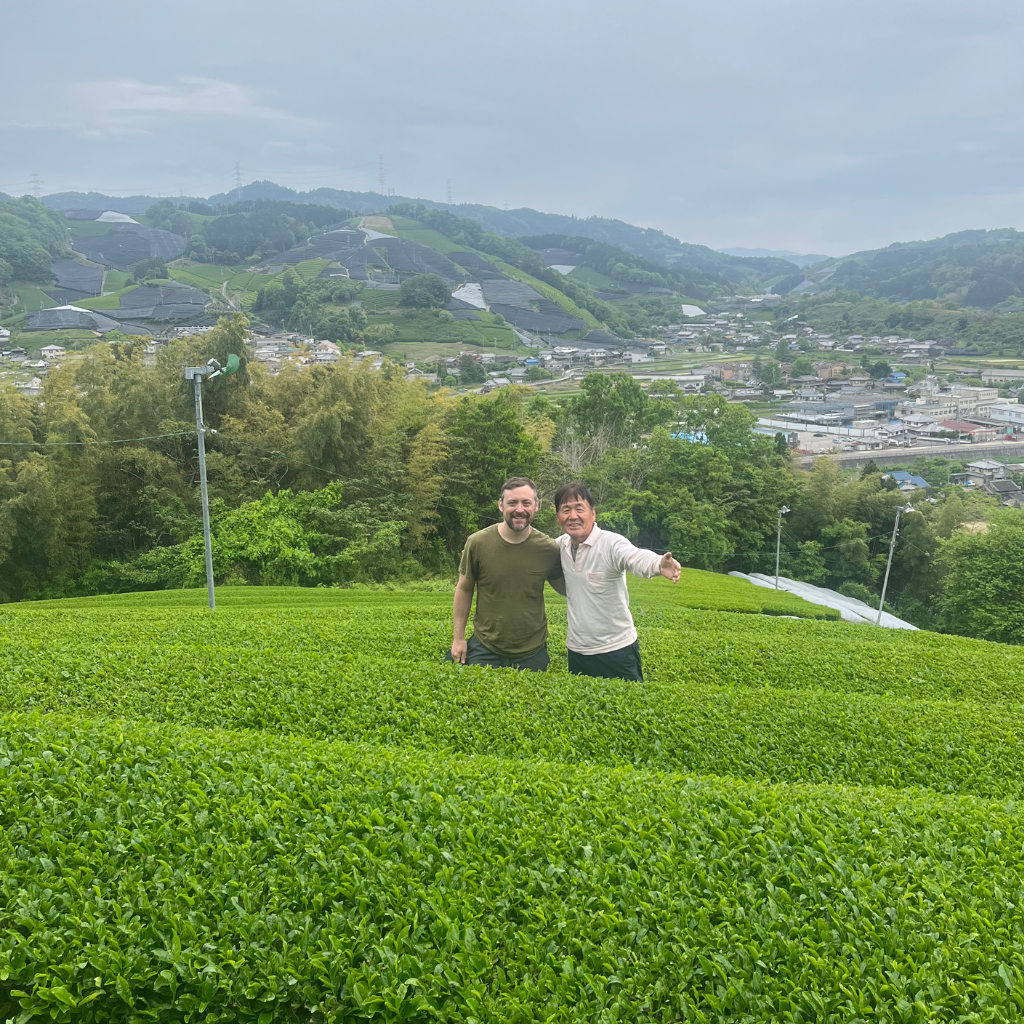 Two individuals stand smiling in lush green tea fields with rolling hills and a quaint village in the background under a cloudy sky.
