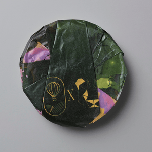 A round, wrapped product, likely a tea cake or puck, wrapped in dark green textured paper with gold foil designs and accents of purple and green, set against a solid gray background.