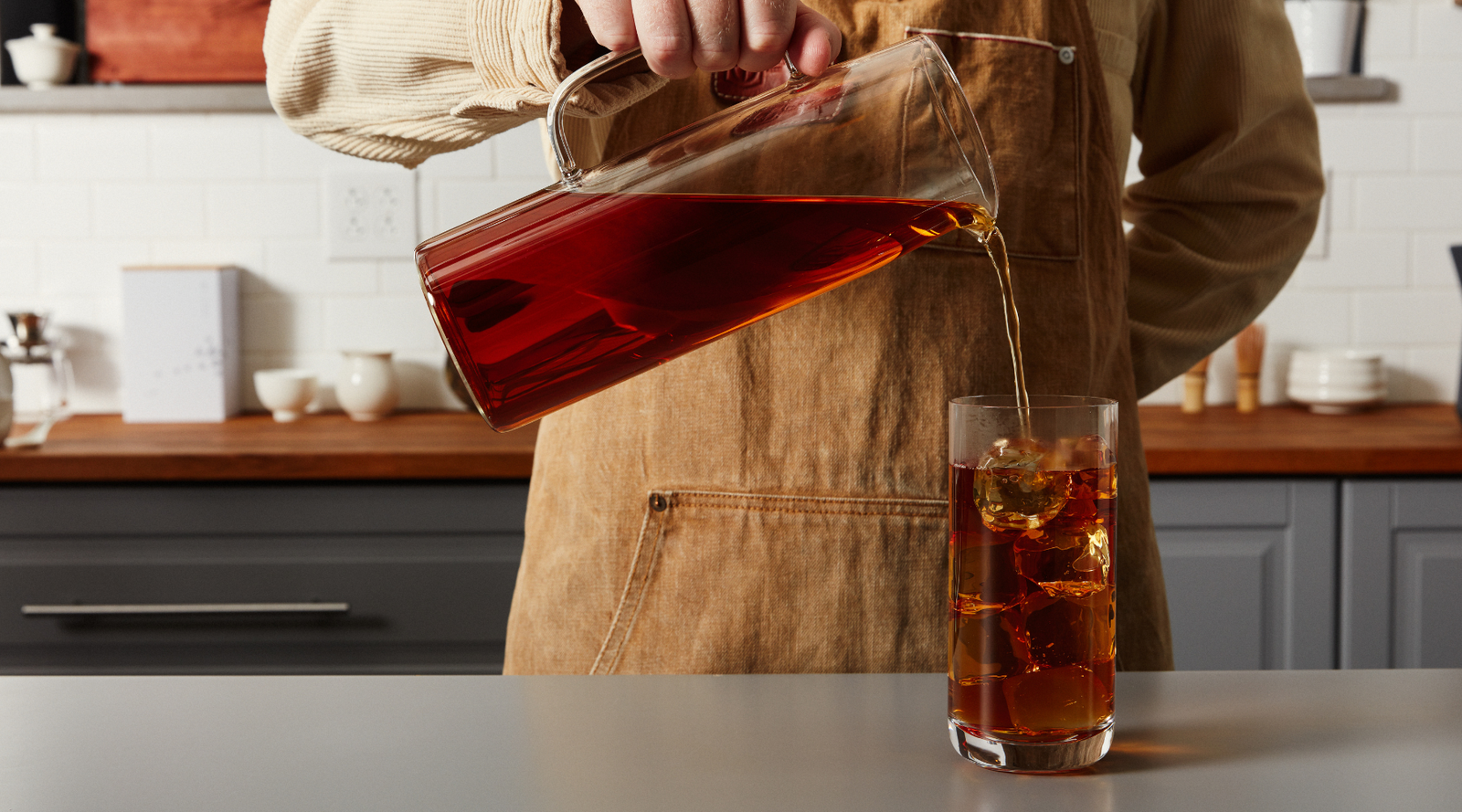 Person pouring iced black tea into a glass on a kitchen counter.