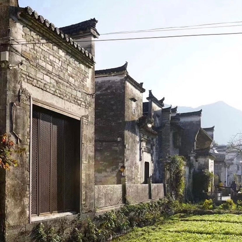 A row of traditional stone houses with ornate roofs under clear skies. Shadows suggest a serene, early morning setting, with mountains in the background.