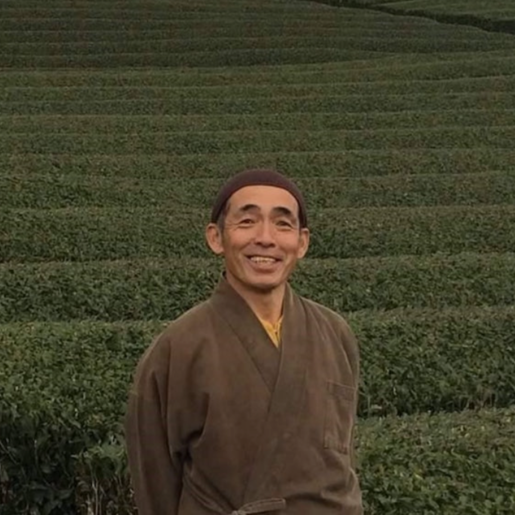 A smiling man in traditional attire stands in front of lush, terraced green fields. The overcast sky adds a serene and peaceful atmosphere.