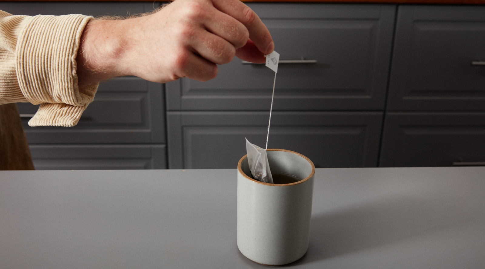 Person placing a tea bag into a mug on a kitchen counter.