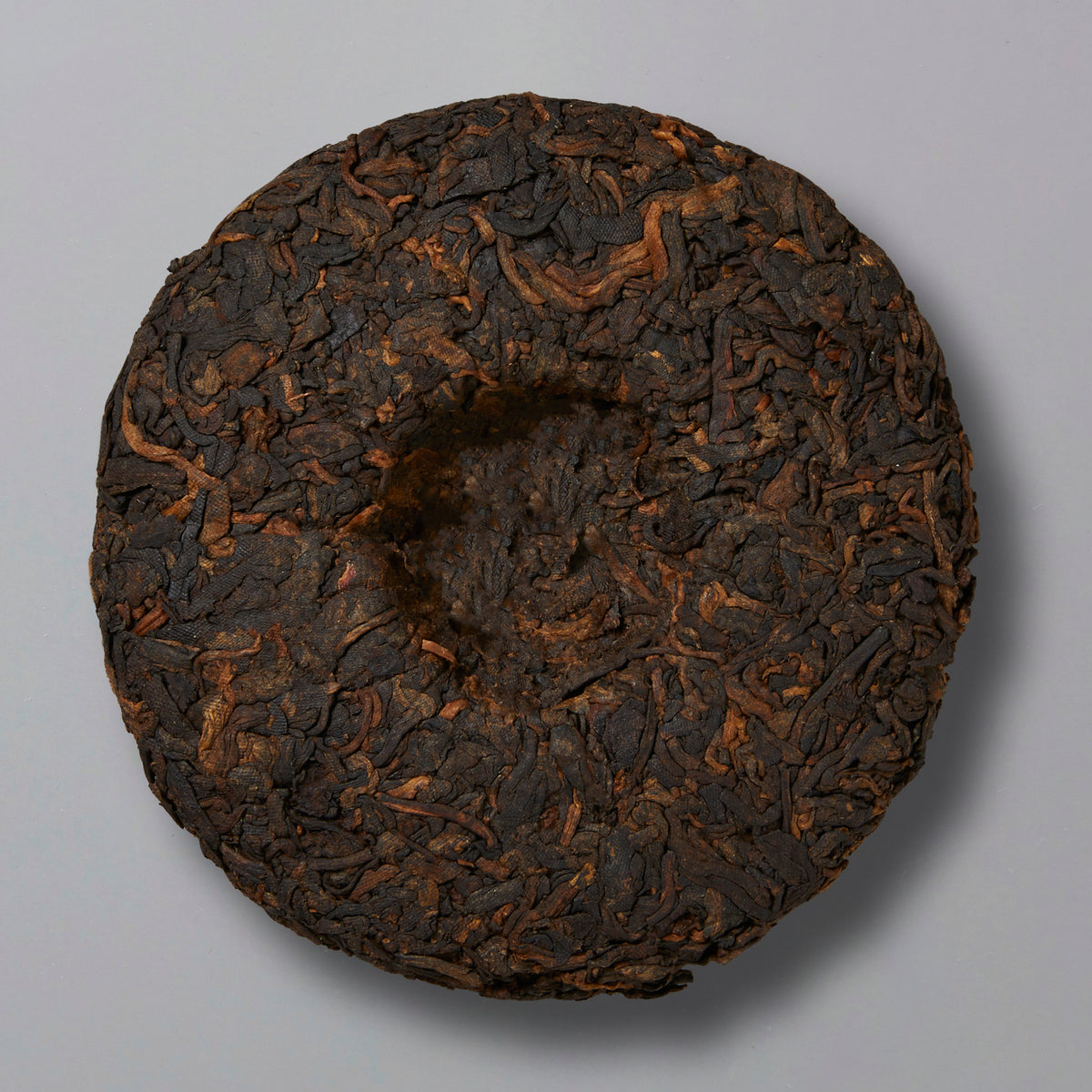 A round, compressed tea cake with various shades of dark brown and black leaves, creating a textured surface, placed on a plain light gray background.