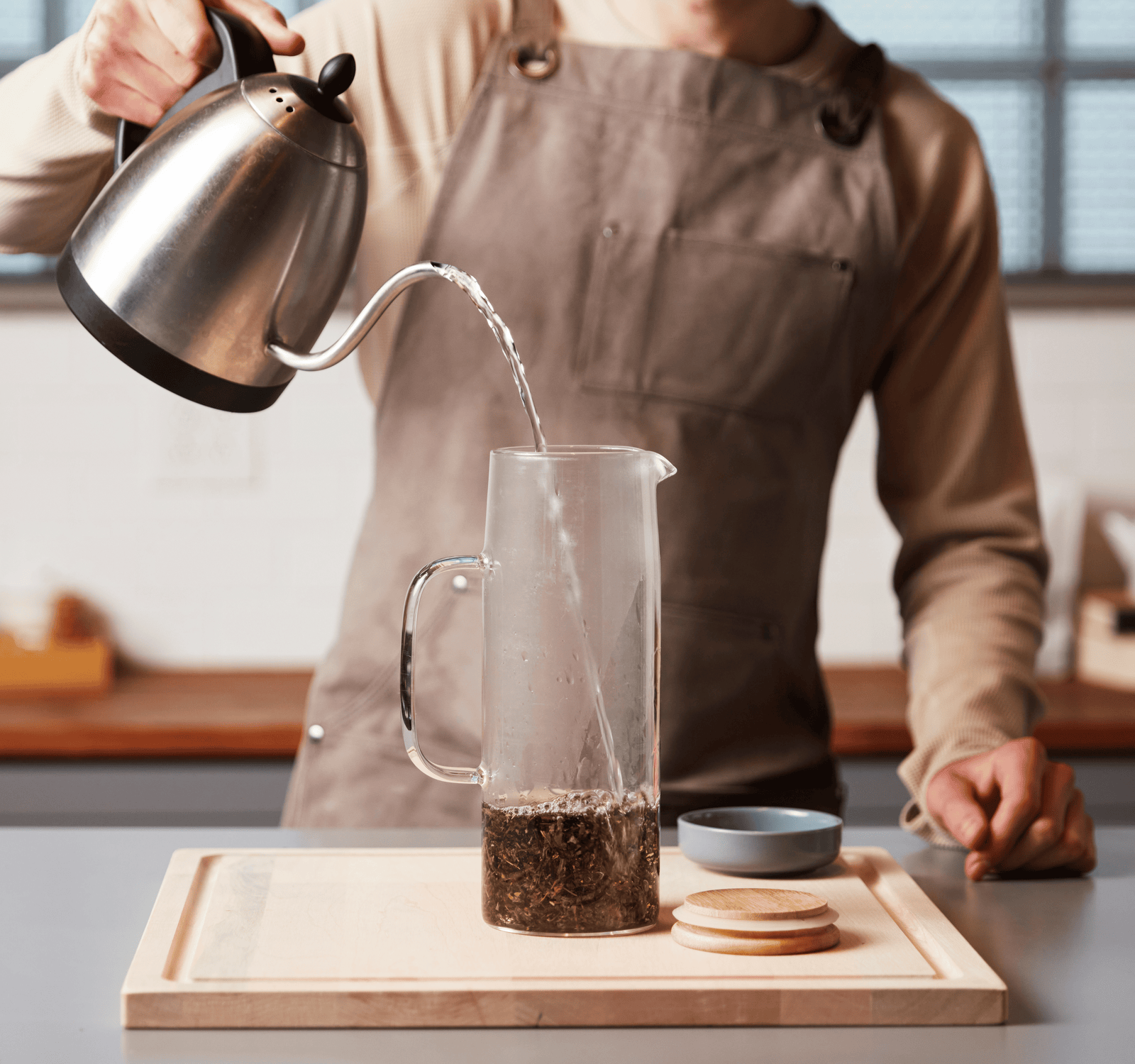 Person pouring hot water into a glass pitcher with tea leaves on a kitchen counter.