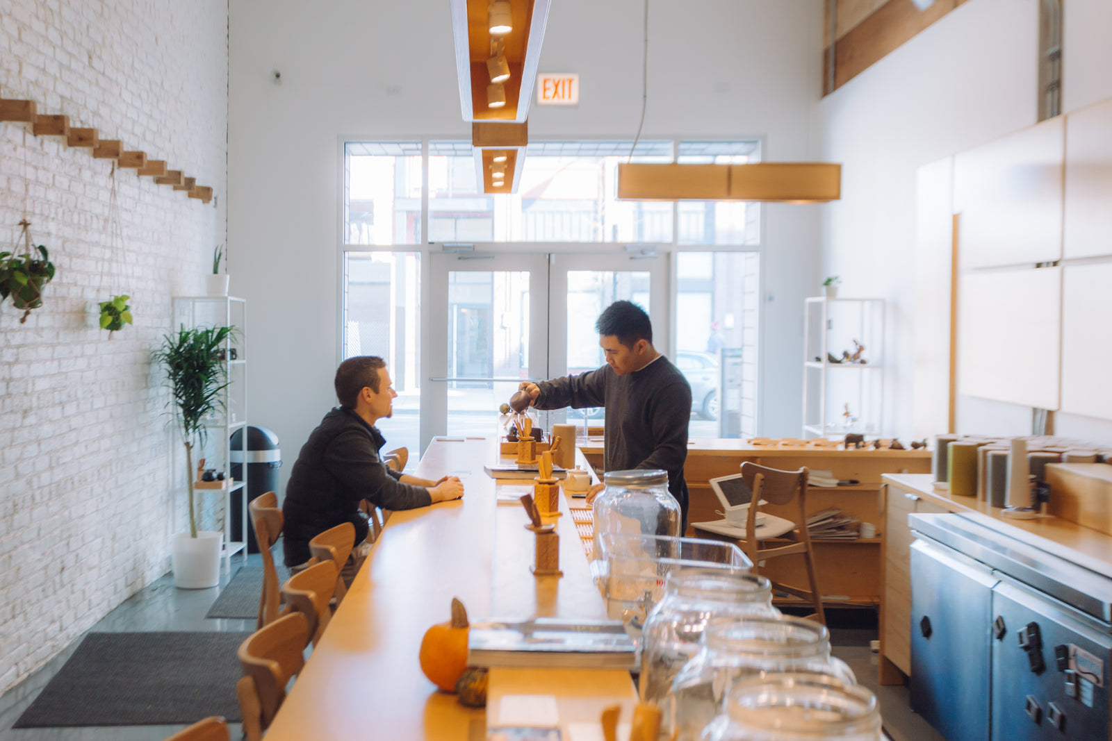 A barista serves tea to a seated customer in a bright, modern cafe with wooden decor and potted plants. The atmosphere is calm and inviting.