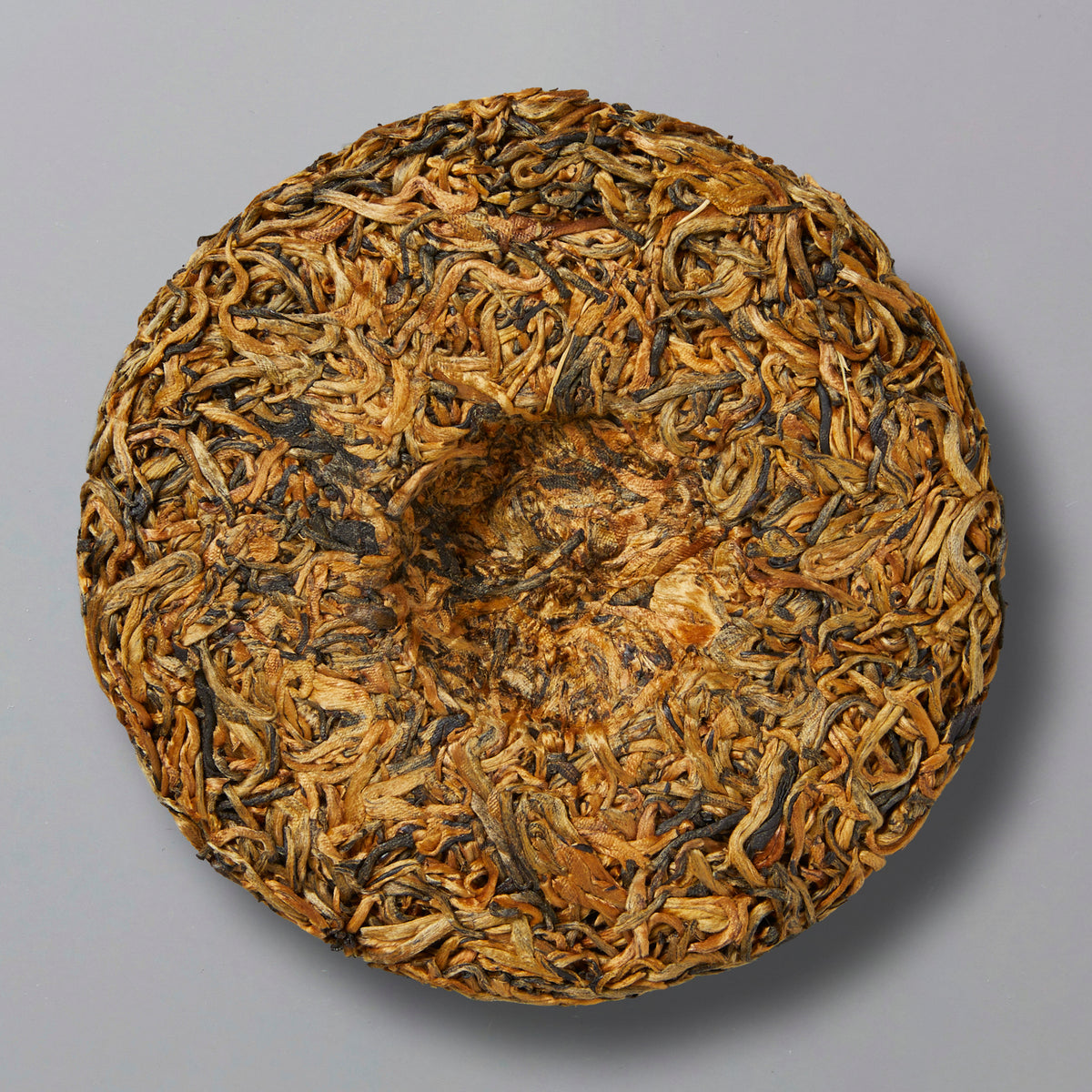 A round disc of tightly packed, dried golden bud black tea leaves, resembling a cake, sits on a plain gray background, conveying a sense of natural texture.