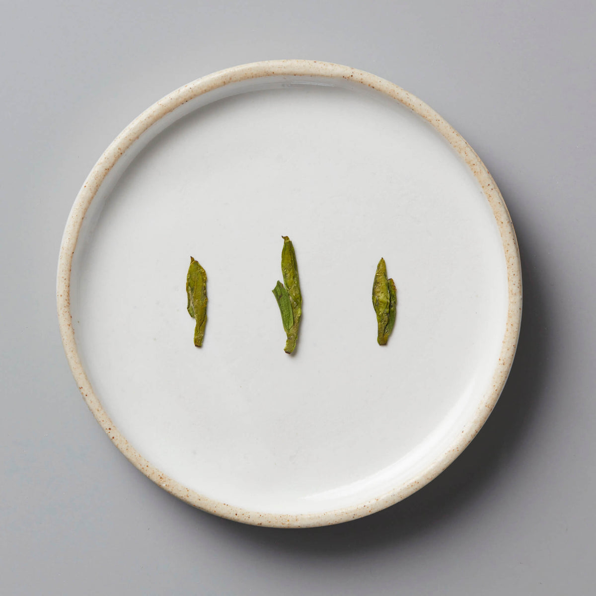 Session Longjing green tea leaves displayed on white ceramic plate, showing characteristic flat pressed dragonwell shape