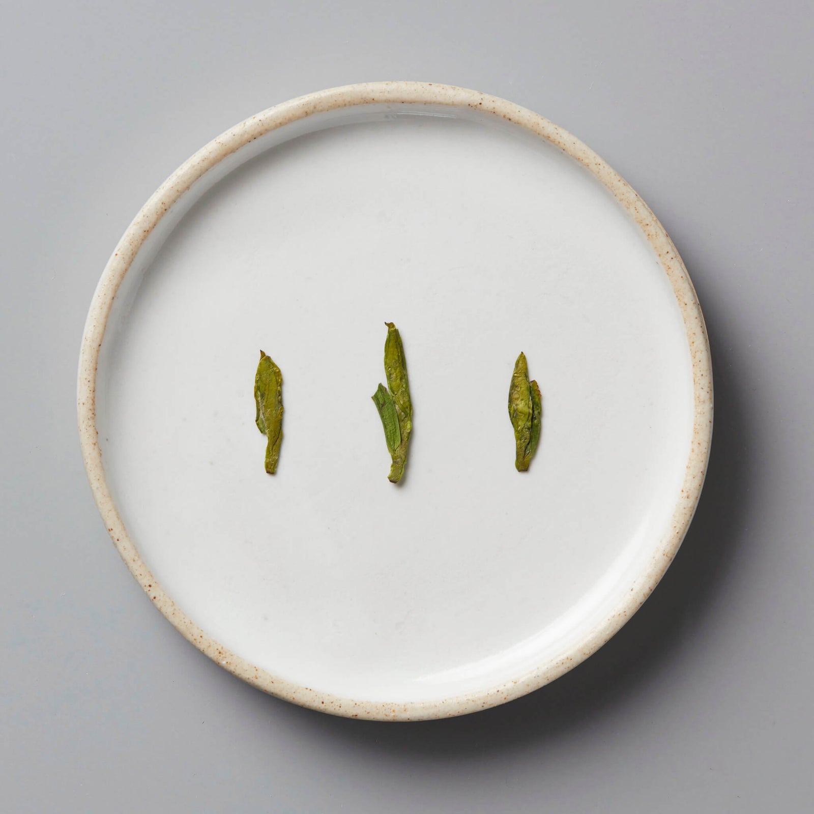 Session Longjing green tea leaves displayed on white ceramic plate, showing characteristic flat pressed dragonwell shape