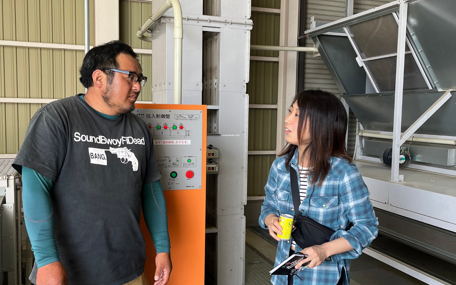 Two people standing in a industrial setting, one holding a yellow cup.