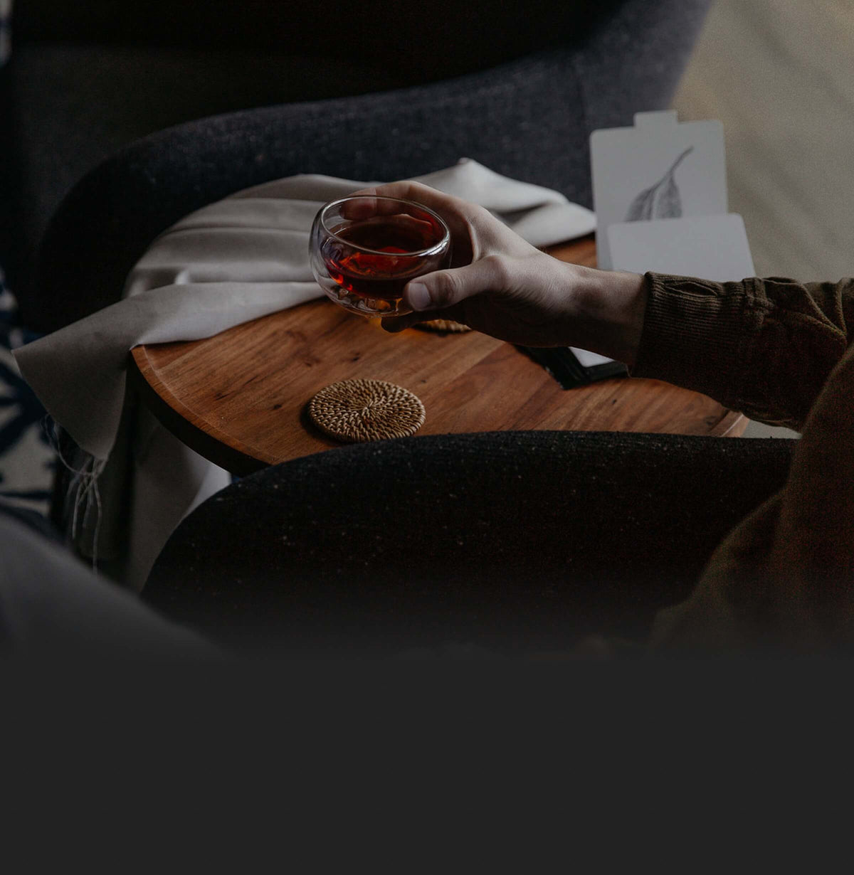 Hand holding a glass cup of tea over a wooden table in a calm, dim setting.