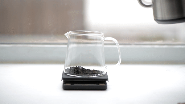 Tea kettle pouring hot water into a mini boli with black tea. The background is well lit and minimalist.
