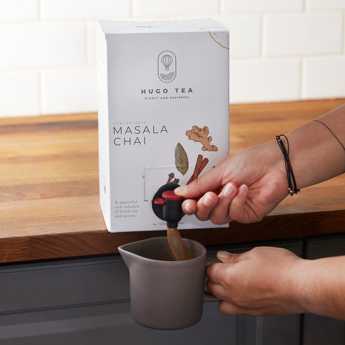 Hands pouring Hugo Tea Masala Chai concentrate from box dispenser into gray ceramic mug on wooden countertop