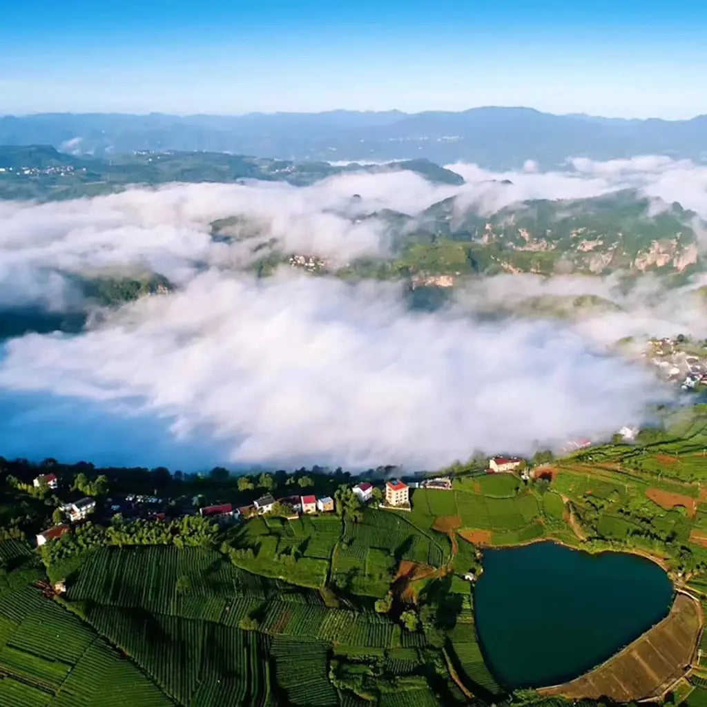 Aerial view of lush green fields with a heart-shaped lake, small houses, and thick white clouds rolling over distant hills under a clear blue sky.