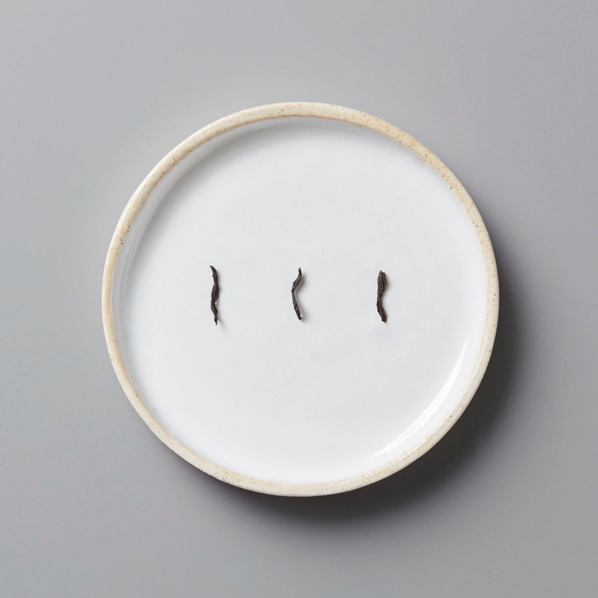 Three small dried black tea leaves on a white ceramic plate with a beige rim on a gray background