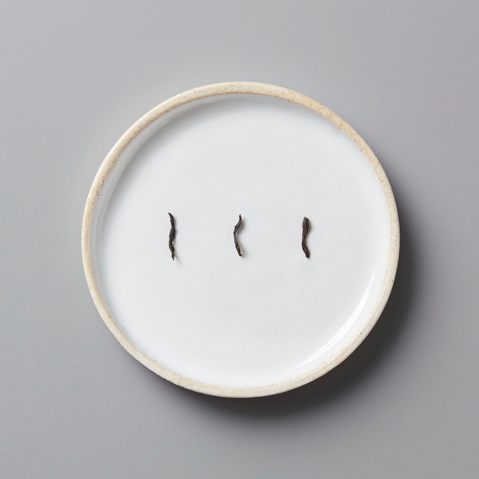 Three small dried black tea leaves on a white ceramic plate with a beige rim on a gray background