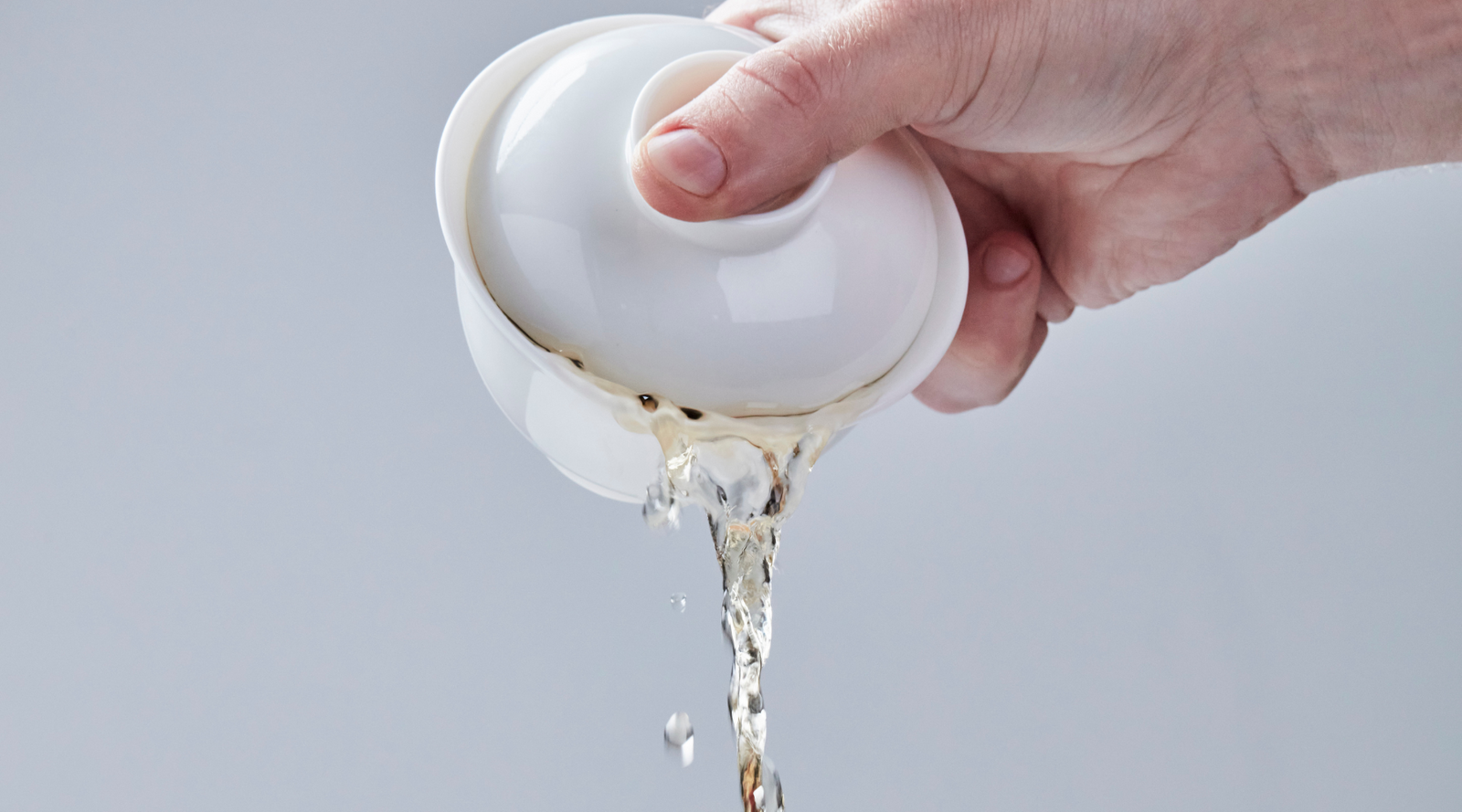 A hand holding a gaiwan is tilted, pouring out brewed tea against a soft gray background. The scene conveys motion and a sense of action.