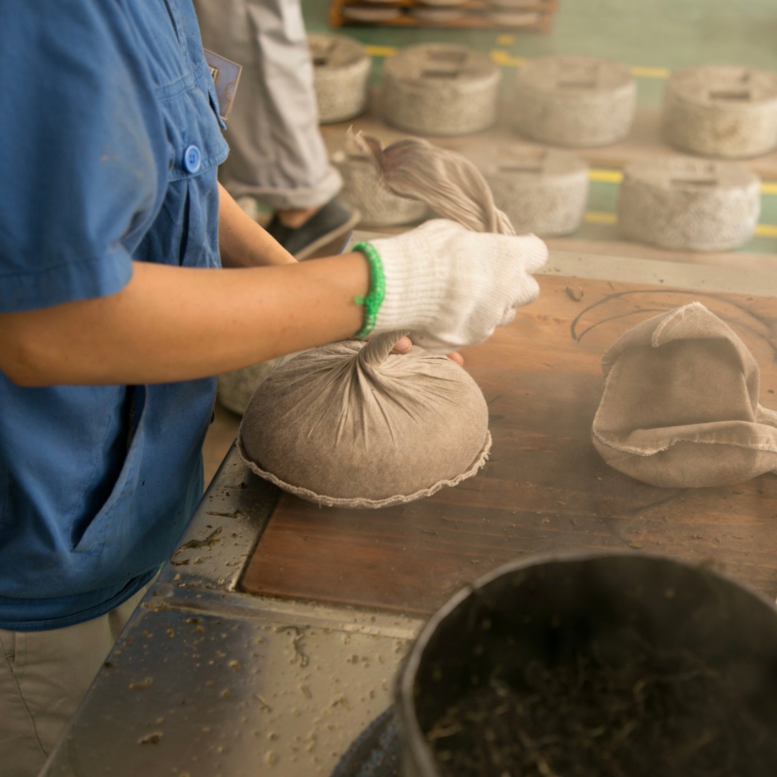 Person in blue shirt and gloves tying a fabric bag filled with plucked tea leaves, set in a workshop with stone grinders in the background. Industrial process.