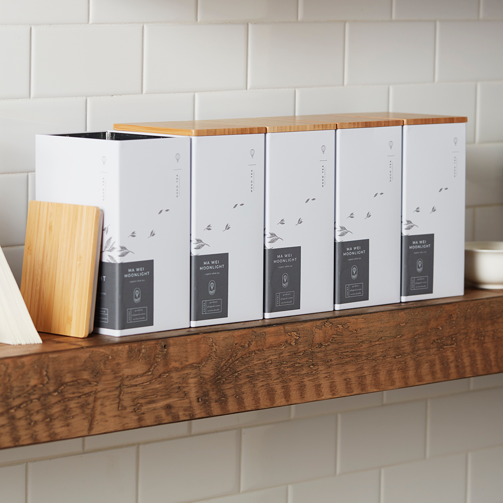 Four white tea tins with bamboo lids labeled "Ma Wei Moonlight" sit neatly on a wooden shelf against a white tiled wall, conveying a minimalist and elegant tone.