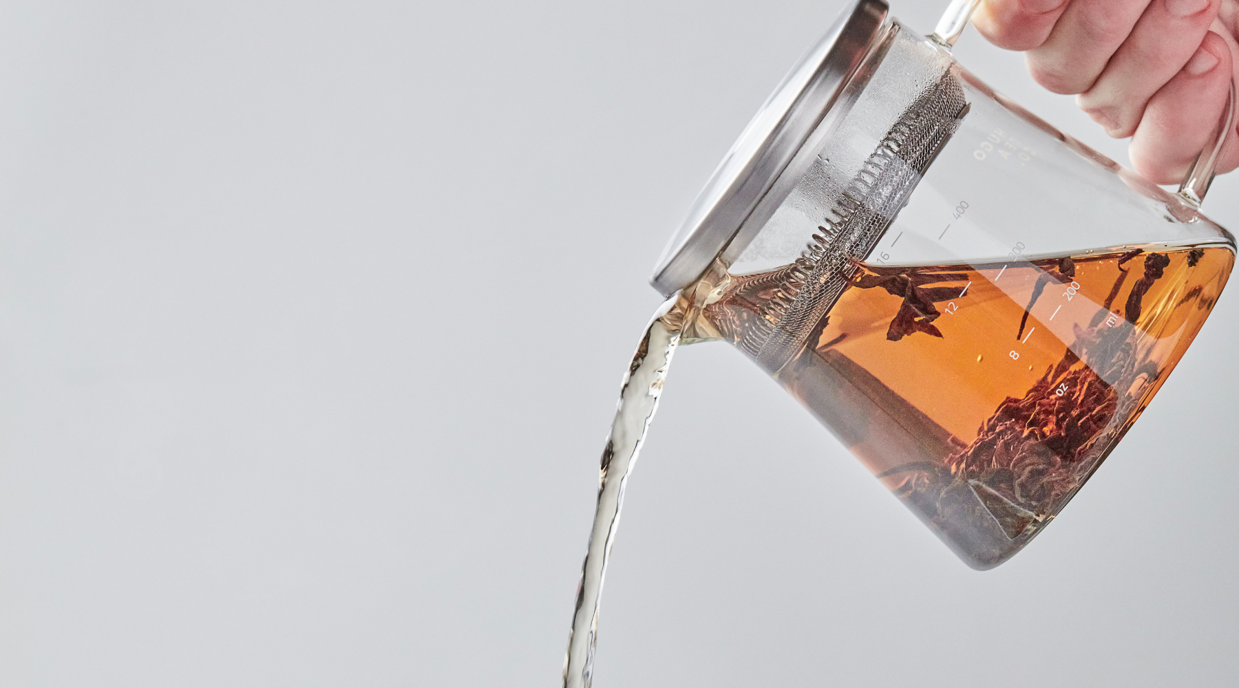 A hand pours amber-colored tea from a glass teapot with a metal filter, against a light gray background, conveying a sense of calm and refreshment.
