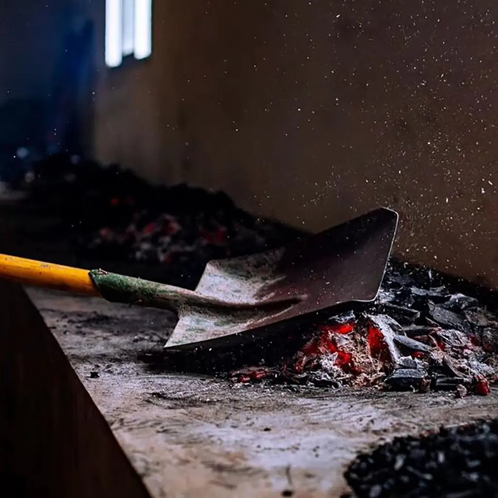 A shovel with a yellow handle stirs glowing red coals and ash on a dark surface. Dust particles float in the air, creating a gritty, rustic atmosphere.