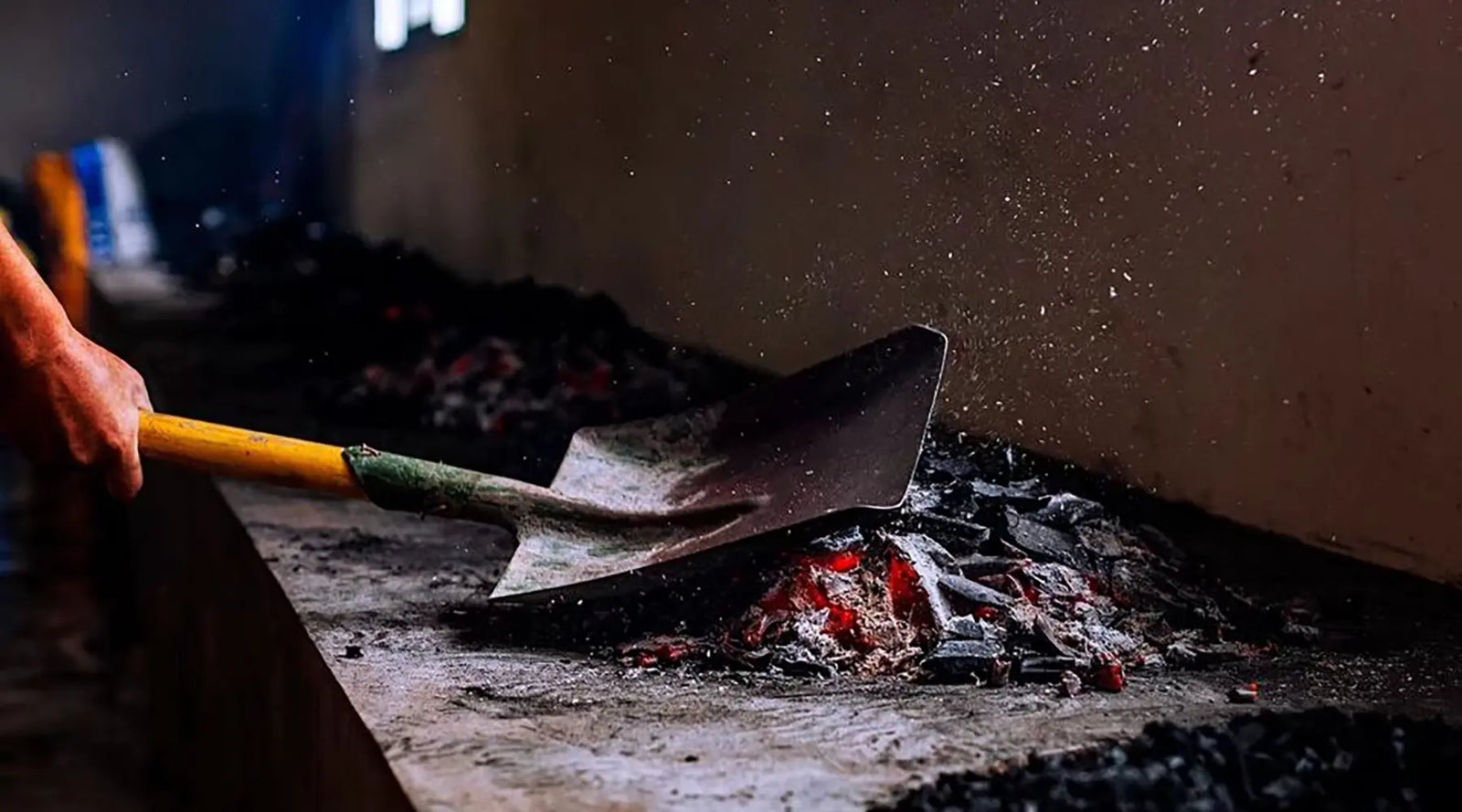 A person uses a shovel to move glowing embers and ash in a dimly lit room, suggesting hard, manual labor. Particles float in the air, creating a gritty atmosphere.