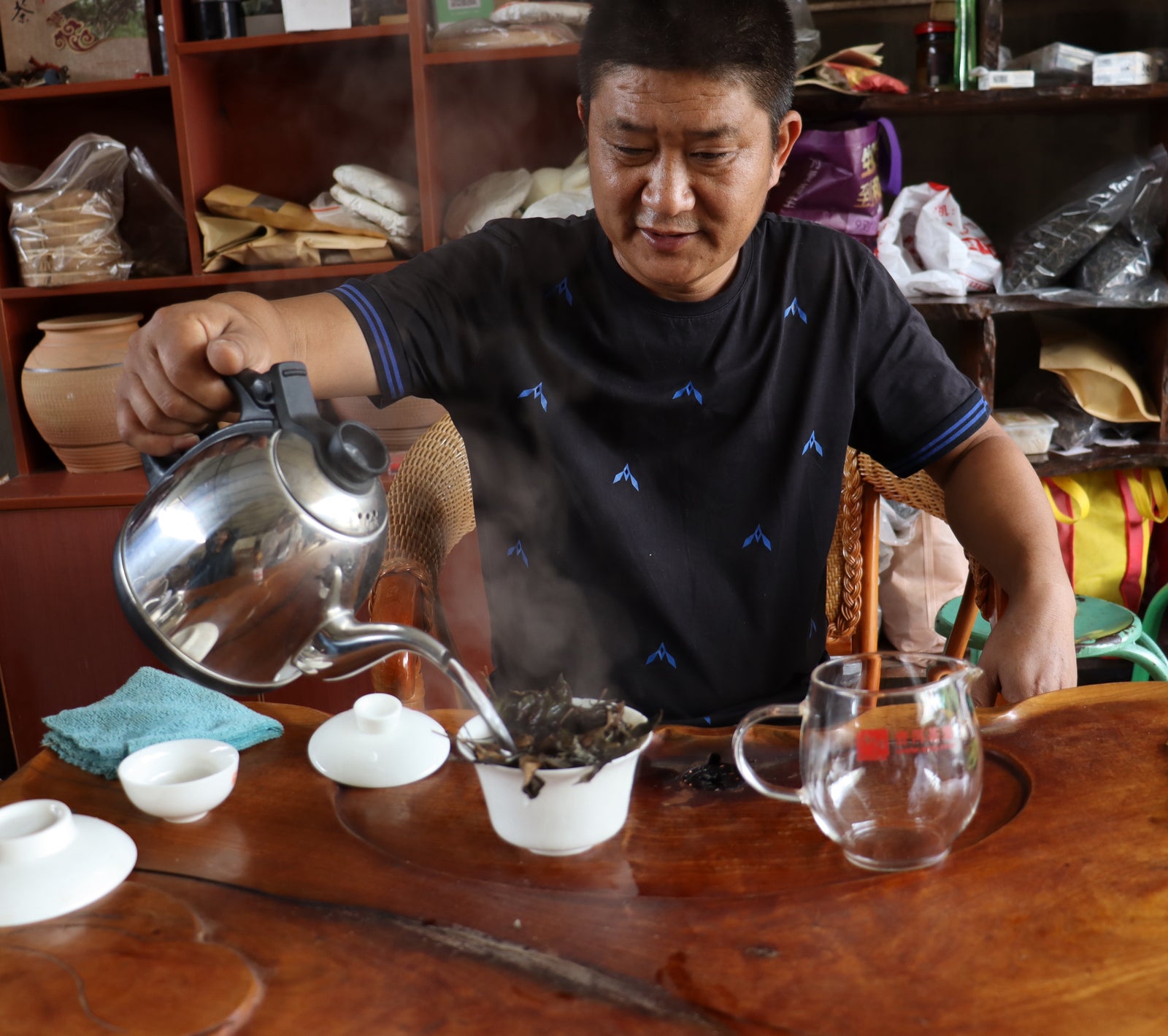 Man pours hot water from a kettle over tea leaves in a teapot, seated at a wooden table. Shelves in the background display various packaged goods.