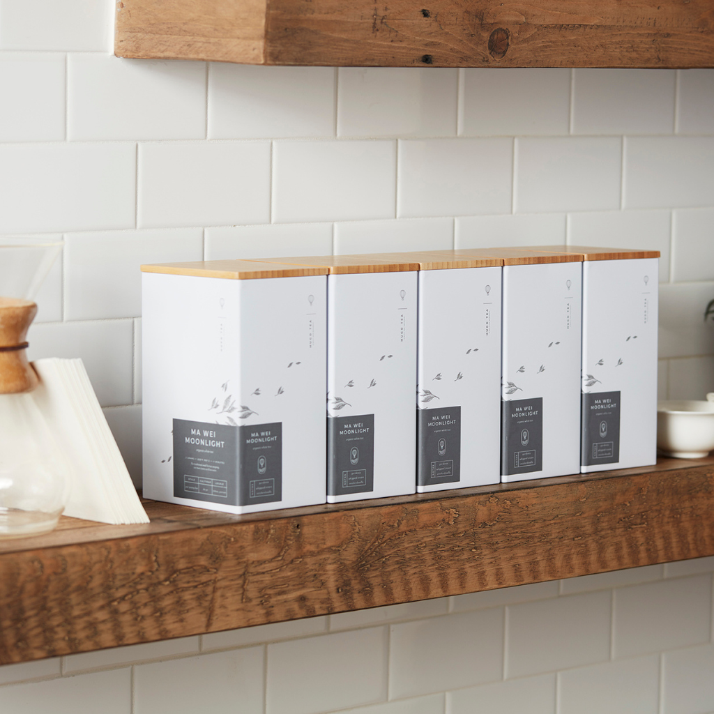 Five neatly arranged white tea boxes with wooden lids sit on a rustic wooden shelf against white tiled walls, creating a clean and minimalist look.