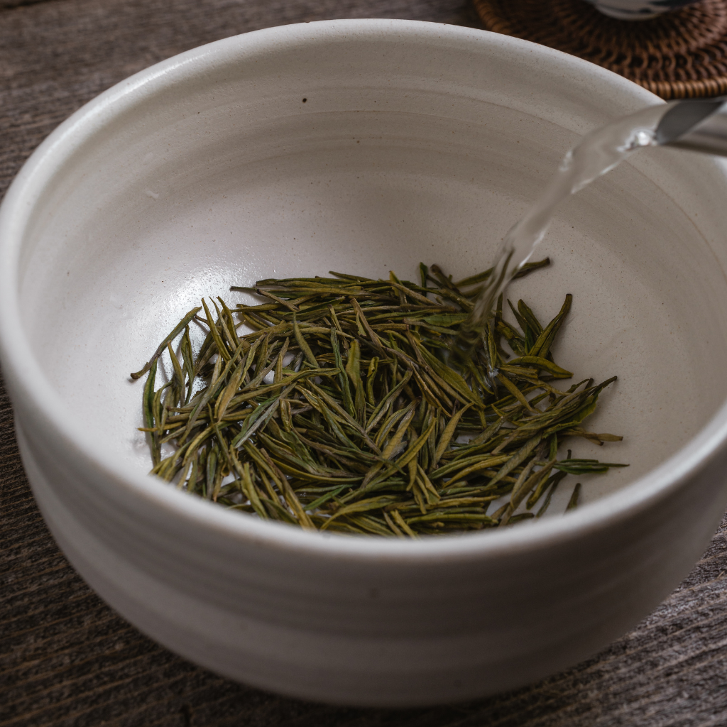 White bowl with green tea leaves being poured into it on a wooden surface.