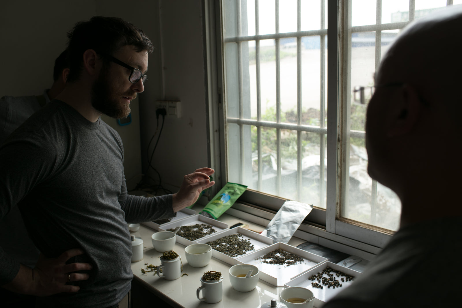 A man in a gray shirt gestures while discussing different tea samples on a table. Several cups and trays with tea leaves sit by a window, creating a thoughtful ambiance.