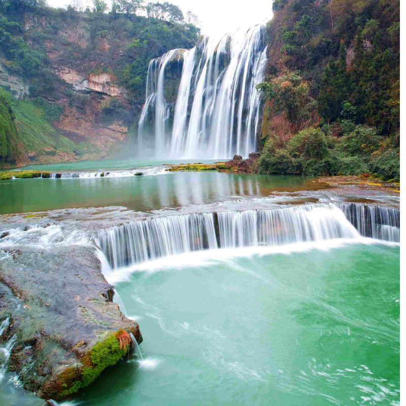 A majestic waterfall cascading over a lush, green cliff into a serene turquoise pool. The scene is tranquil, with mist rising and vibrant foliage.