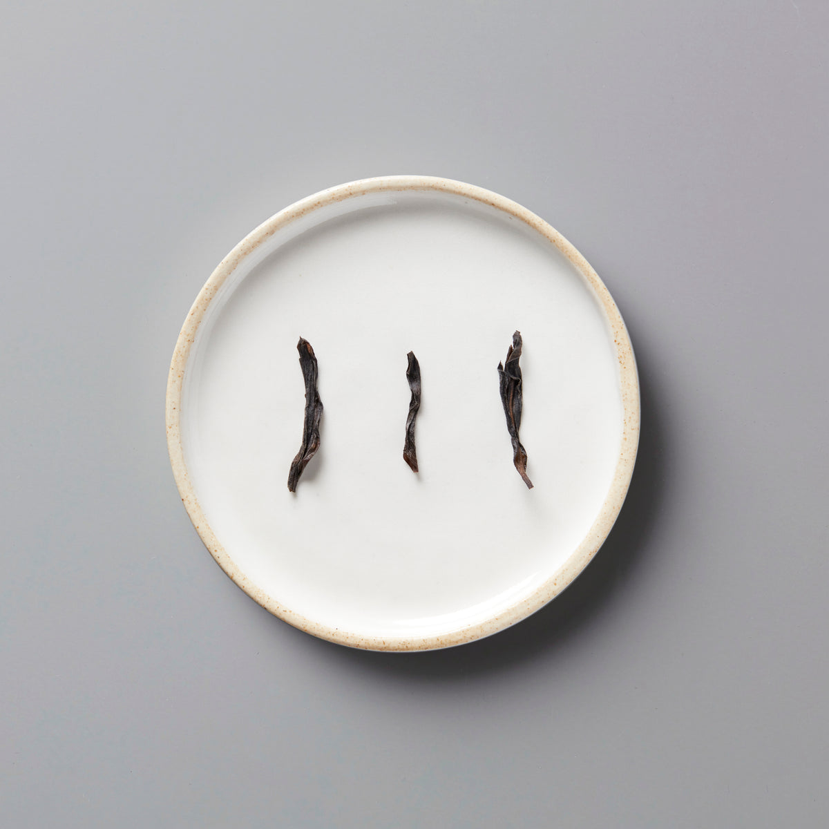 Three twisted dark tea leaves of LINS DUCK dancong oolong arranged on a white ceramic plate with beige rim