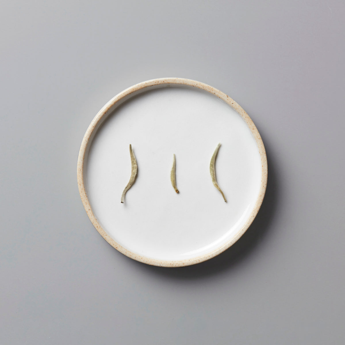 Ma Wei Needles white tea buds displayed on white ceramic plate with beige rim against grey background