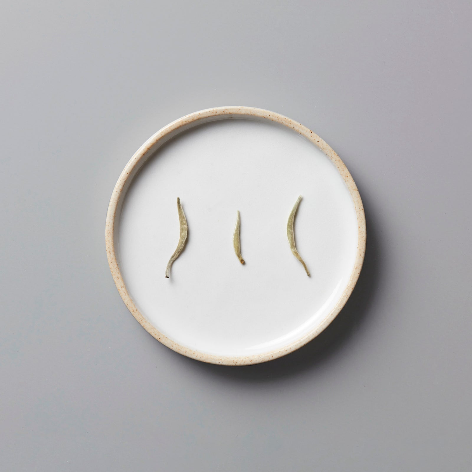 Ma Wei Needles white tea buds displayed on white ceramic plate with beige rim against grey background