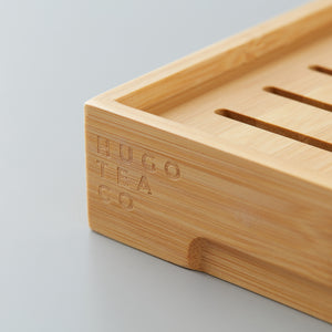 Close-up of the corner of a bamboo TEA BOARD HUGO tea tray with engraved branding on a smooth surface.